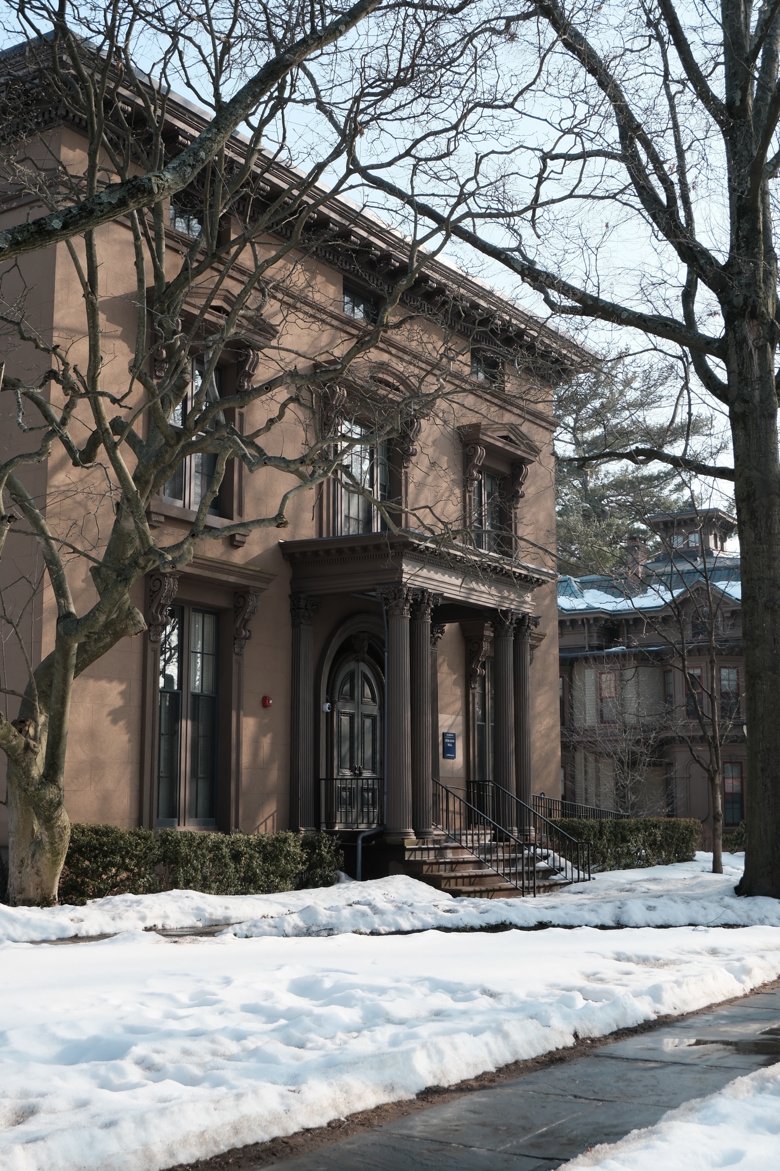 A stucco Italianate house with a columned portico, framed by bare branches above fresh snow