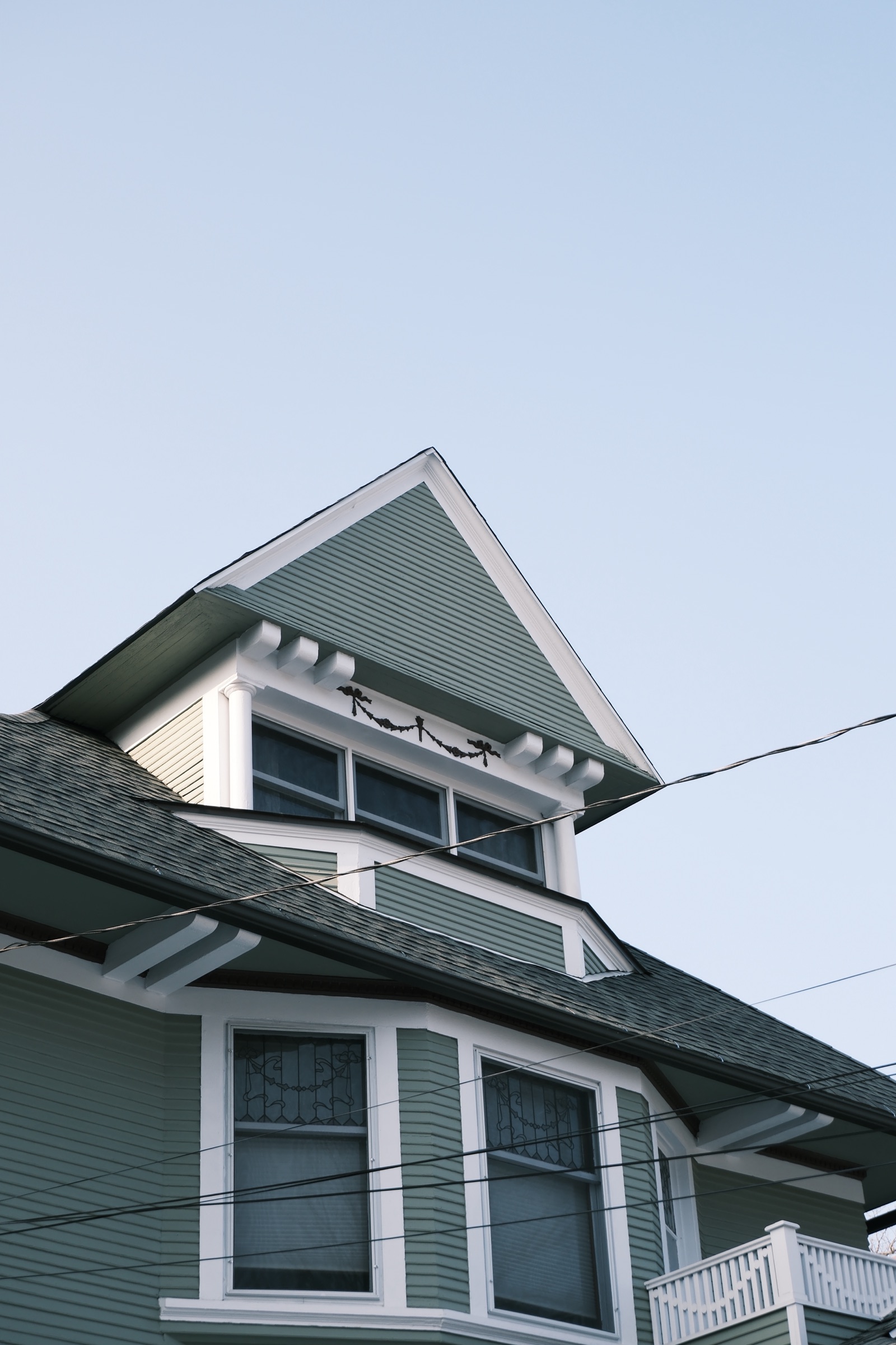 The gable and dormer of a pale green Victorian house against a clear winter sky, crossed by power lines