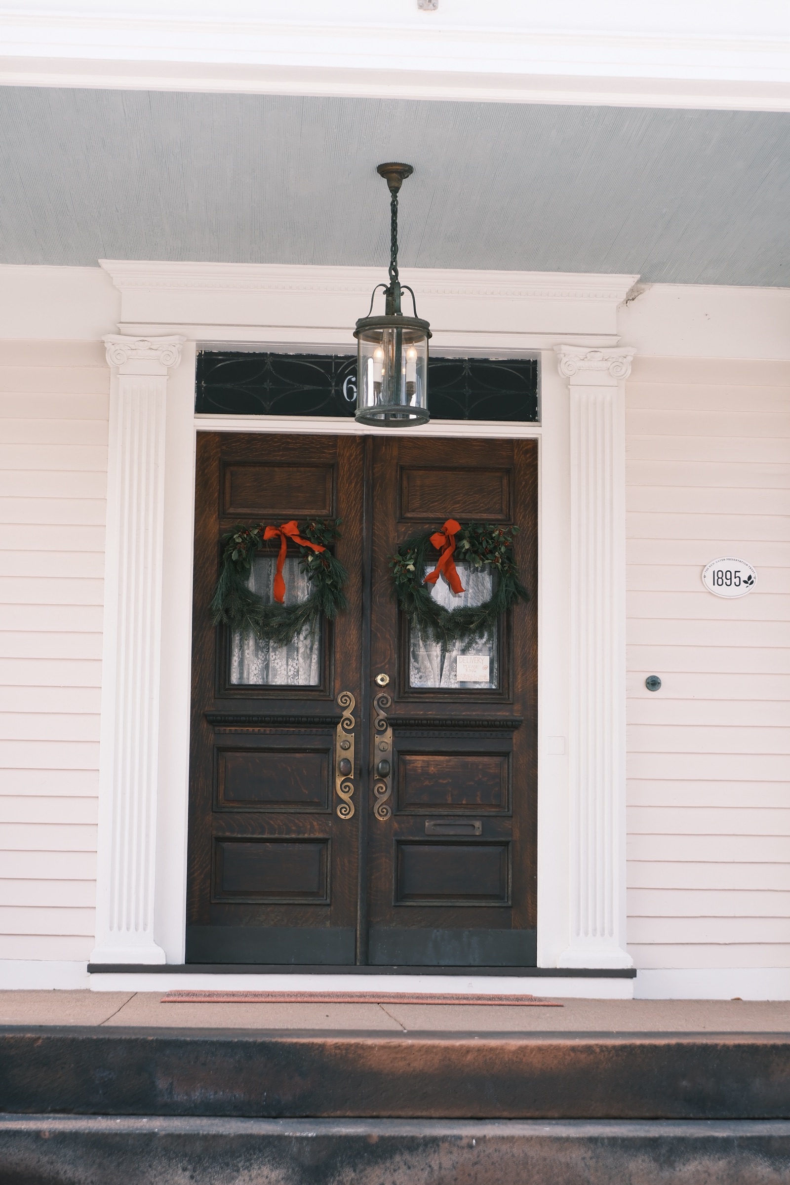 Dark wooden double doors hung with holiday wreaths, lit by a lantern on a white porch