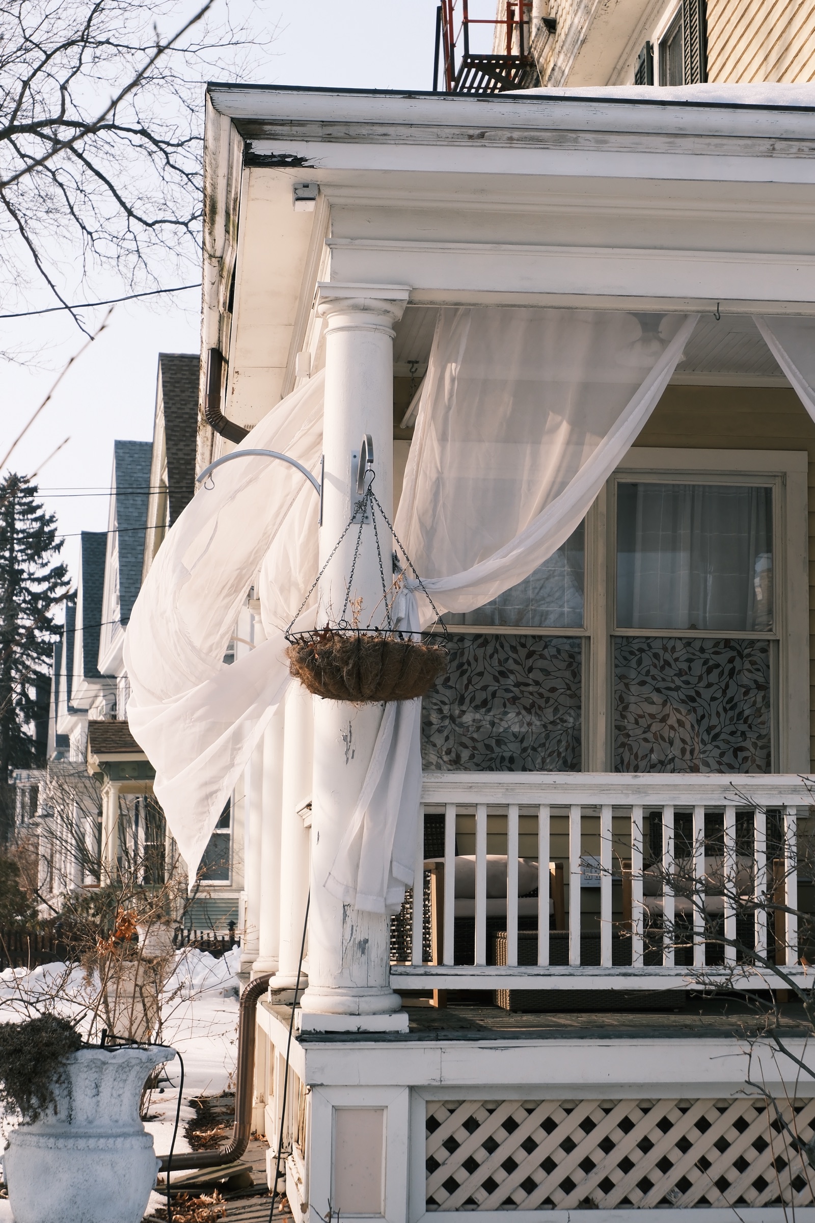 White curtains blowing from a porch column beside an empty hanging planter