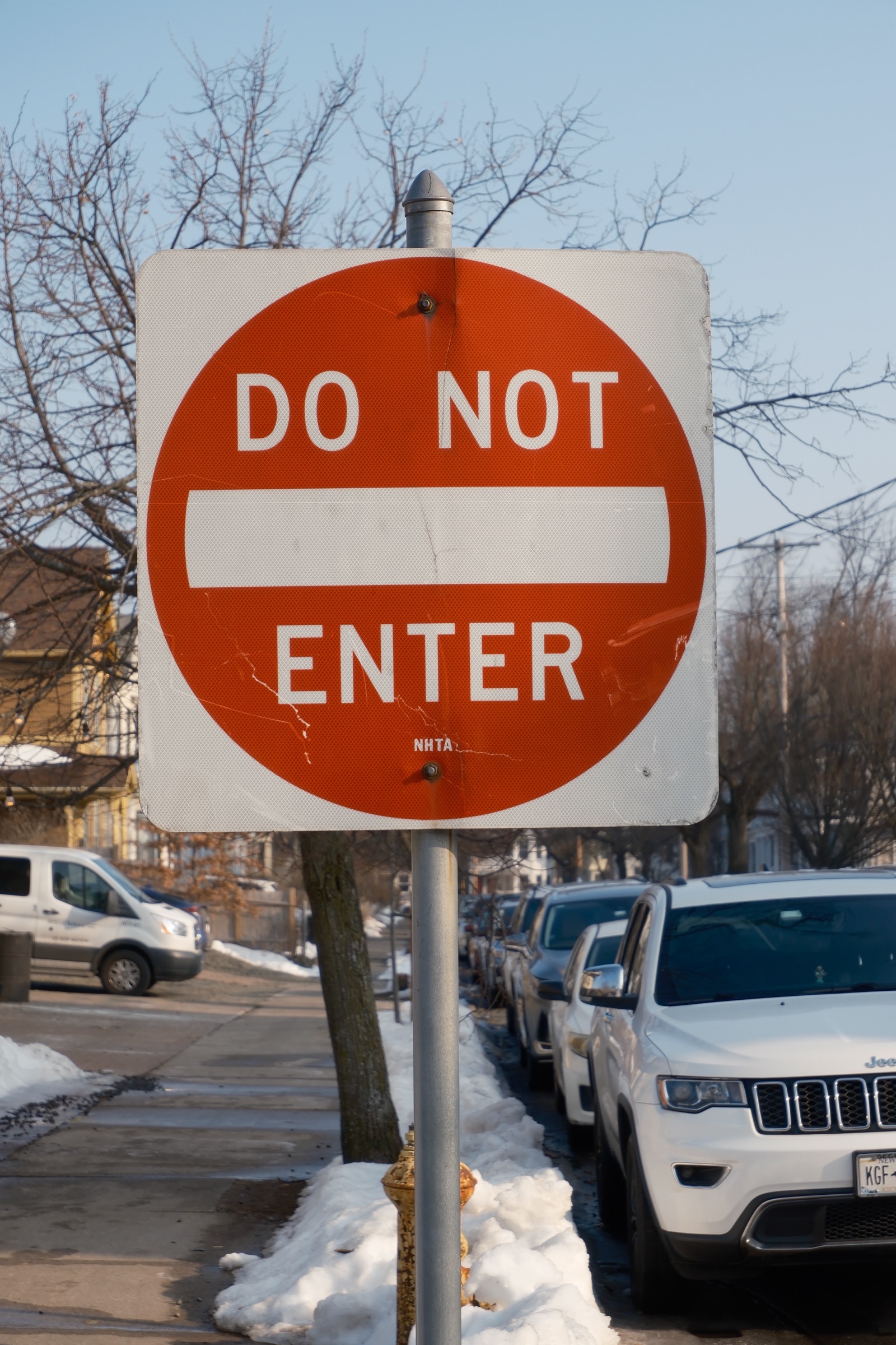 A weathered DO NOT ENTER sign standing above a row of cars parked along a snowy street