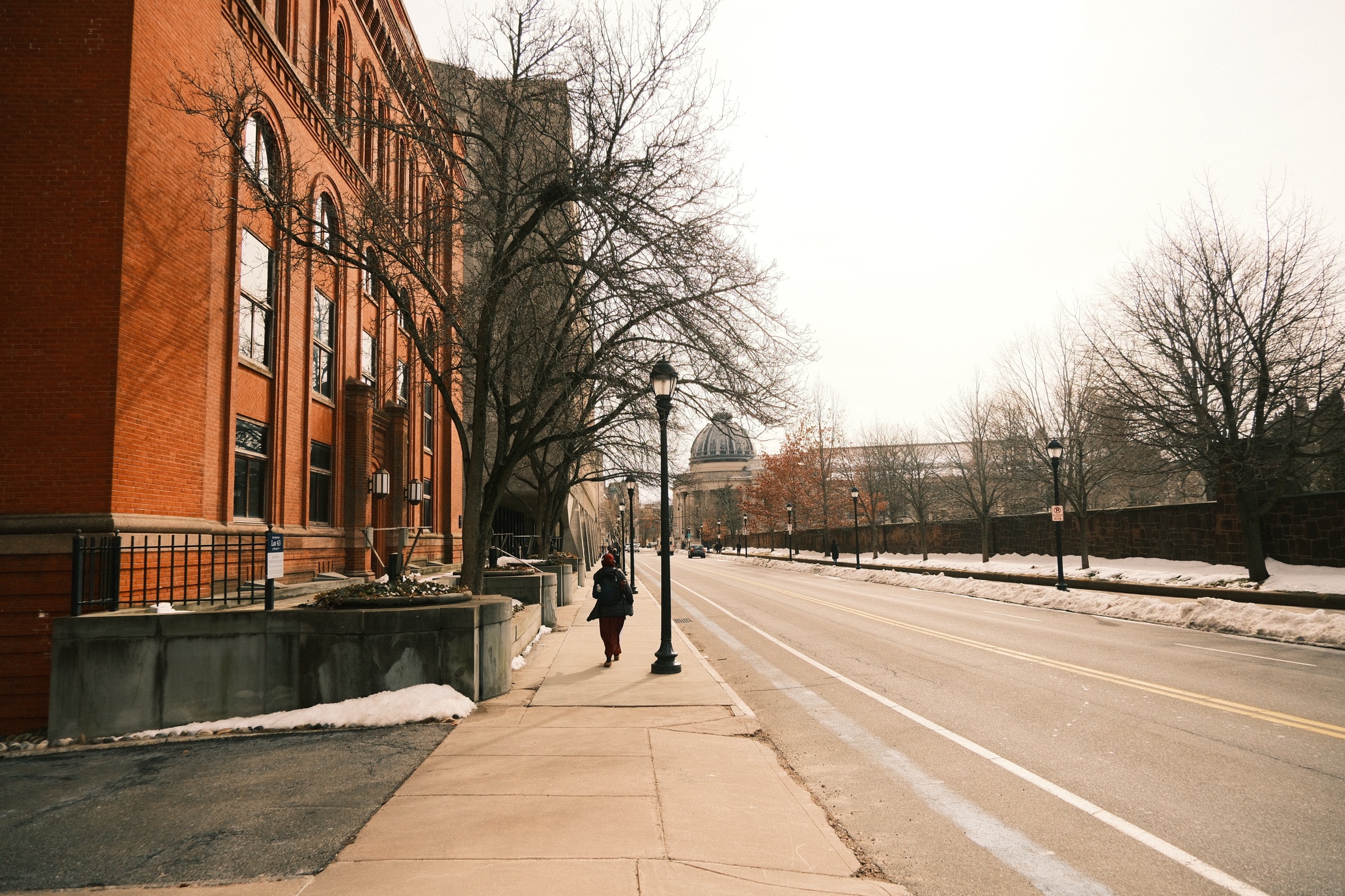 A lone figure walking a wide, empty street past a red brick building toward a distant dome